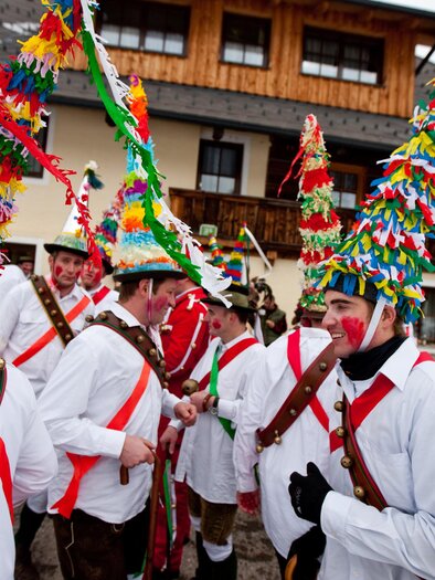 Eine Gruppe von Männern in traditionellen kostümierten Kleidern mit bunten Hüten feiert vor einem Holzhaus. Sie tragen weiße Hemden und sind mit bunten Bändern geschmückt. | © Tv Murau