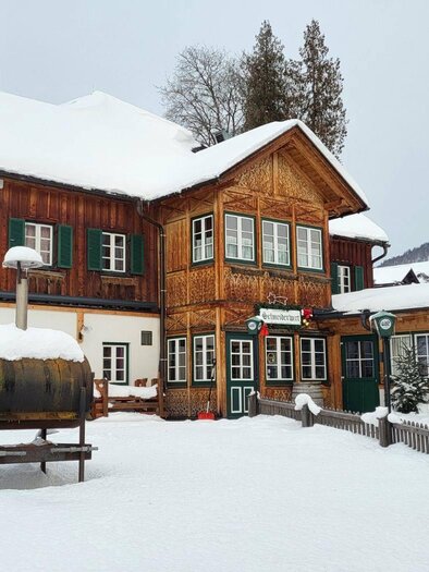 The traditionally "Ausseer" house of the Schneiderwirt tavern in the snow, surrounded by the winter landscape of Altaussee. The sky is overcast and the atmosphere feels calm and peaceful. | © Petra Kirchschlager