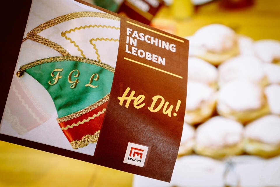 Ein Flyer für Fasching in Leoben mit bunten Motiven und dem Text „He Du!“. Im Hintergrund sind traditionelle Krapfen sichtbar. | ©  Foto Freisinger