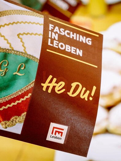 A flyer for Carnival in Leoben with colorful motifs and the text "Hey You!". Traditional donuts are visible in the background. | ©  Foto Freisinger