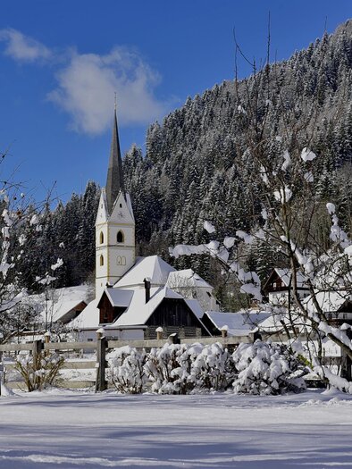 Eine verschneite Winterlandschaft mit einer Kirche und malerischen Bergen im Hintergrund. Die Bäume sind mit Schnee bedeckt, und der Himmel ist blau mit einigen Wolken.
