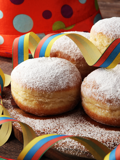 Delicious donuts with powdered sugar are lying on a wooden plate. Colorful party ribbons and a hat are decorated around the donuts. | © Fasching Ausseerland Füllbild (c) canva