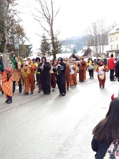 Die Salinenmusikkapelle führt den traditionellen Faschingsumzug mit den verkleideten Gruppen in bunten Kostümen durch die Straßen von Altaussee an. Im Hintergrund sind verschneite Bäume und Häuser der Gemeinde am Fuße des Losers zu sehen. | © Kirchschlager Petra