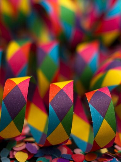 Bunte Konfetti-Bänder mit einem Muster aus Rauten liegen auf einer Fläche aus glitzerndem Konfetti. Die Farben strahlen Freude und Feierlaune aus. | © streamer-g