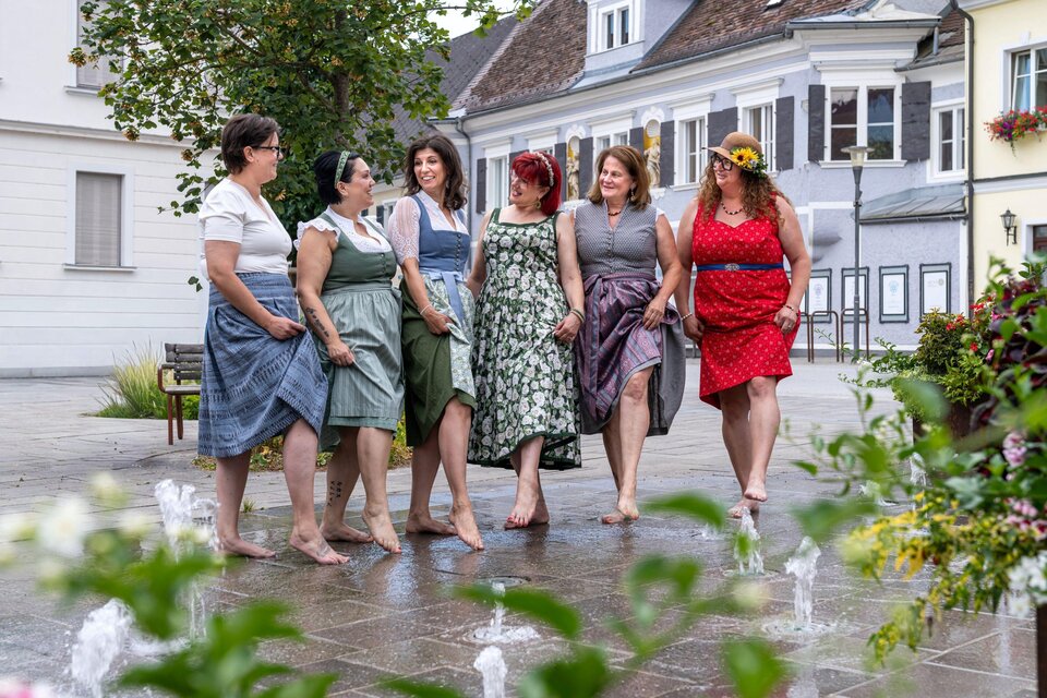 Sechs Frauen in bunten Kleidern stehen barfuß und lächeln in einer Stadt. Im Hintergrund sind Häuser und Pflanzen zu sehen. | © Studio4 Christian Freydl