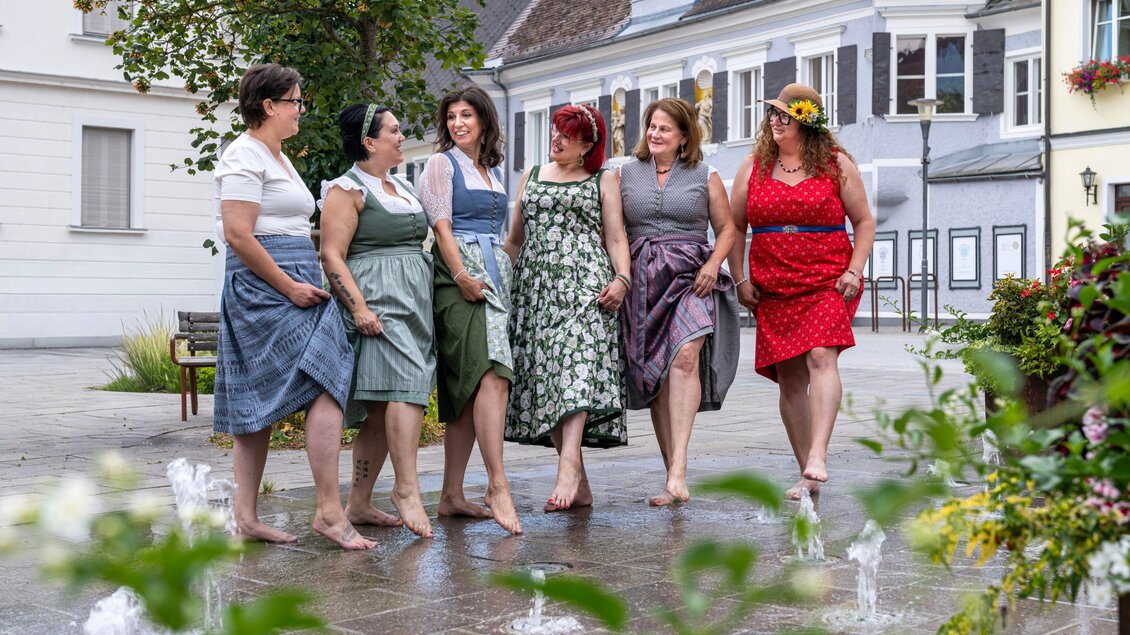 Sechs Frauen in bunten Kleidern stehen barfuß und lächeln in einer Stadt. Im Hintergrund sind Häuser und Pflanzen zu sehen. | © Studio4 Christian Freydl