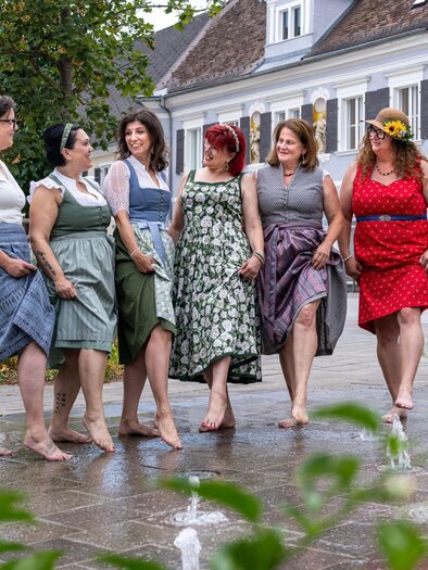 Six women in colorful dresses stand barefoot and smile in a city. In the background, houses and plants can be seen. | © Studio4 Christian Freydl