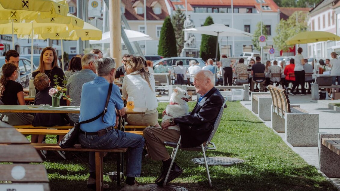 Eine belebte Außenszene mit mehreren Gruppen, die an Tischen sitzen und sich unterhalten. Sonnenschirme und grüne Wiesen sorgen für eine einladende Atmosphäre. | © Anna Zora