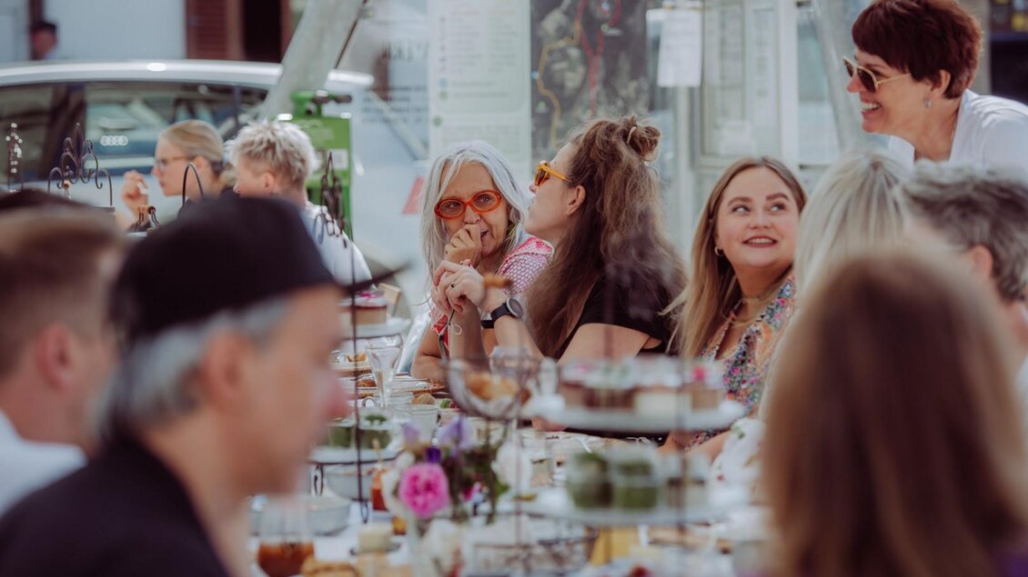 Eine fröhliche Gruppe von Menschen sitzt an einem Tisch und unterhält sich. Der Tisch ist festlich gedeckt und es gibt eine entspannte Atmosphäre. | © Anna Zora