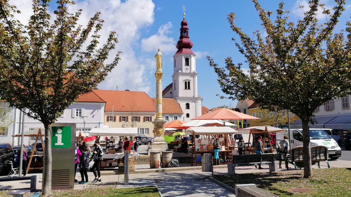 Ein lebhafter Platz mit Ständen und einer Statue im Zentrum. Im Hintergrund ist eine Kirche mit einem markanten Kirchturm zu sehen, umgeben von Bäumen und Besucher. | © Stadtgemeinde Fehring