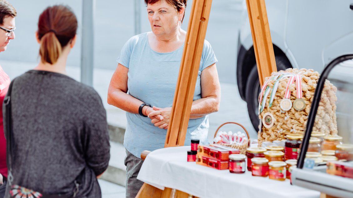 Eine Verkaufsstand mit verschiedenen Marmeladen und Auszeichnungen ist zu sehen. Eine Frau spricht mit zwei Kundinnen und erklärt ihre Produkte. | © Lumikki Photography