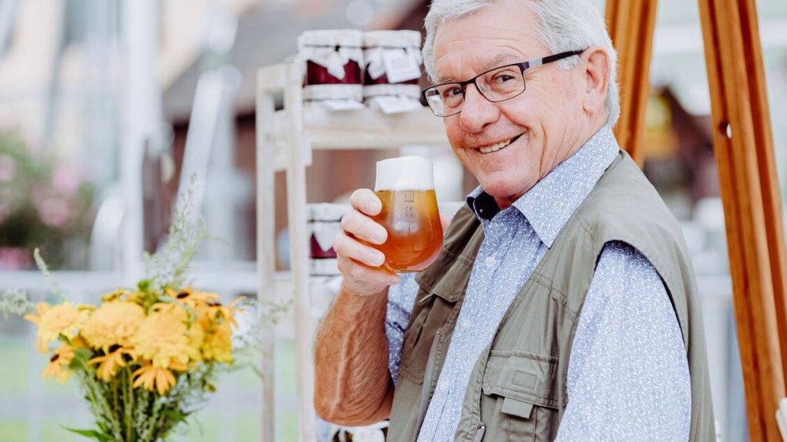 Ein fröhlicher älterer Mann hält ein Bierglas in der Hand und lächelt. Im Hintergrund sind Blumen und ein Regal mit Gläsern zu sehen. | © Lumikki Photography