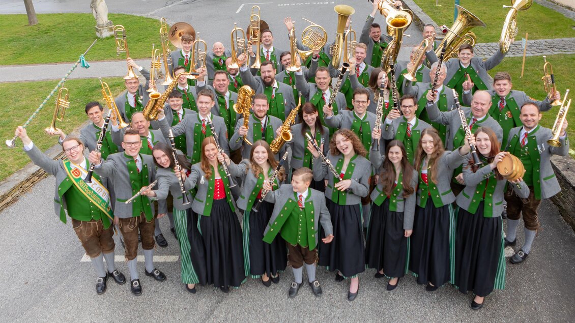 Eine große Musikgruppe steht im Freien und hält ihre Instrumente in die Luft. Die Mitglieder tragen traditionelle Kleidung mit grünen Elementen. | © TMV St. Magdalena a.L.