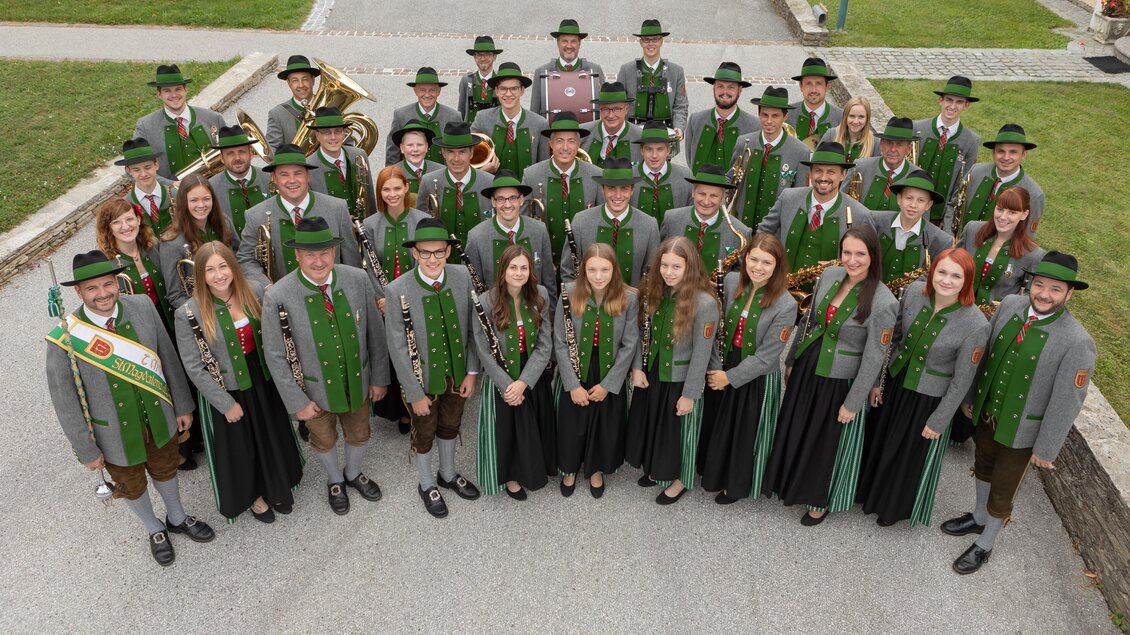 Eine große Gruppe von Menschen in traditionellen Trachten steht auf einem Platz. Sie sind gut gelaunt und posieren gemeinsam für ein Gruppenfoto.
