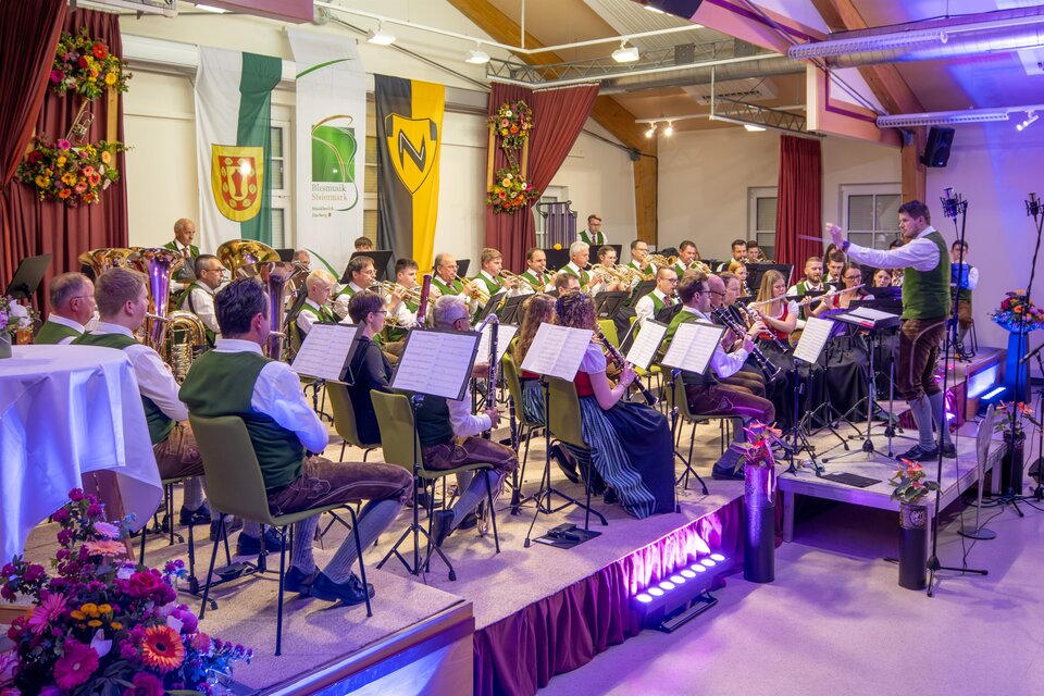 Ein Blasorchester spielt in einem festlich dekorierten Saal. Der Dirigent leitet die Musiker, die in traditionellen Kleidung sitzen. | © Mittlinger