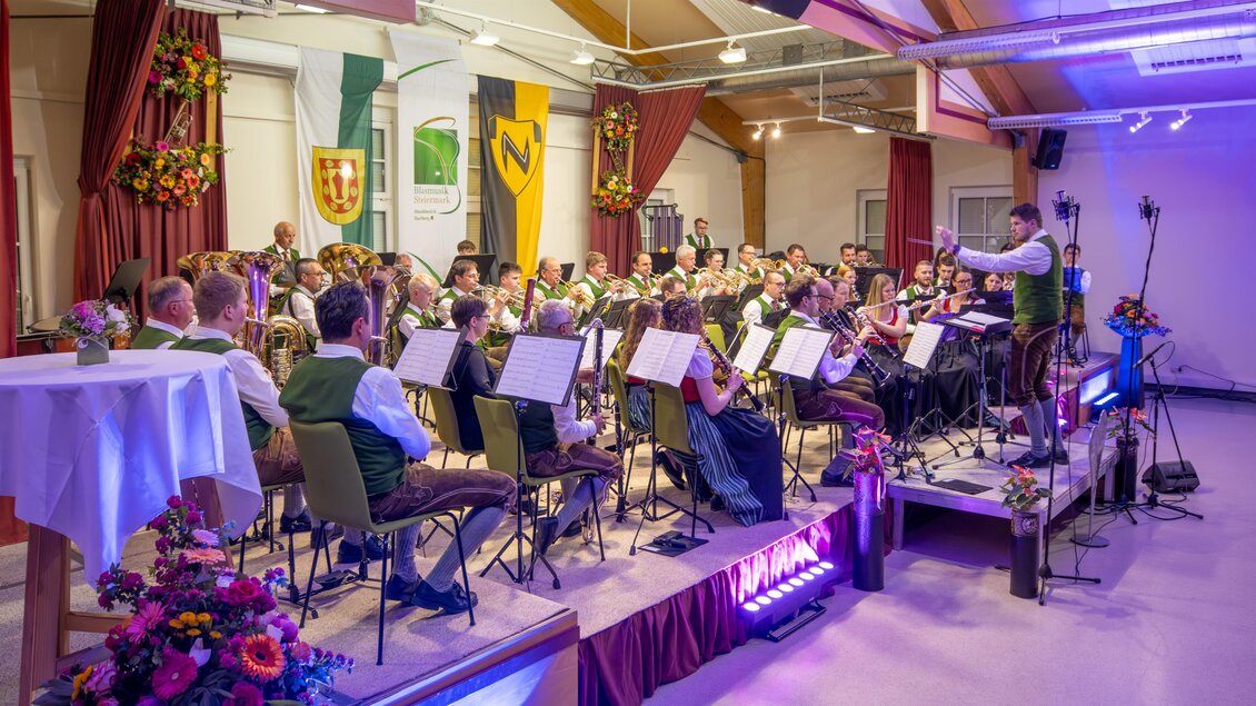 Ein Blasorchester spielt in einem festlich dekorierten Saal. Der Dirigent leitet die Musiker, die in traditionellen Kleidung sitzen. | © Mittlinger