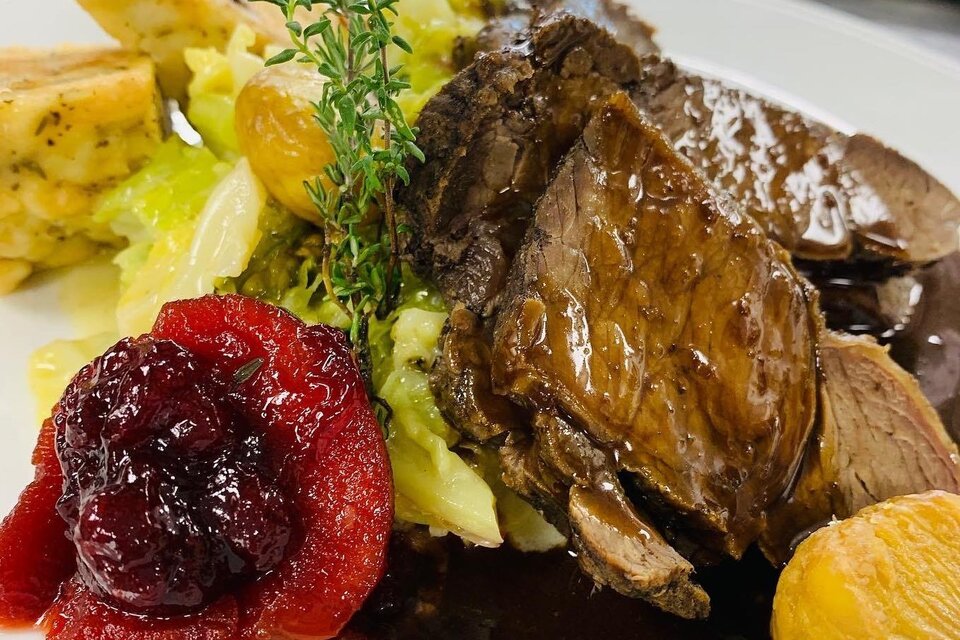 Ein köstlicher Teller mit zartem Fleisch, Beilagen aus Gemüse und Klößen. Das Gericht wird mit Preiselbeeren und einer Kräuterdekoration serviert. | © Wirtshaus Meißl