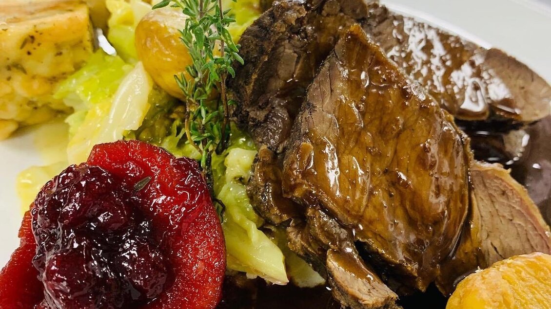Ein köstlicher Teller mit zartem Fleisch, Beilagen aus Gemüse und Klößen. Das Gericht wird mit Preiselbeeren und einer Kräuterdekoration serviert. | © Wirtshaus Meißl