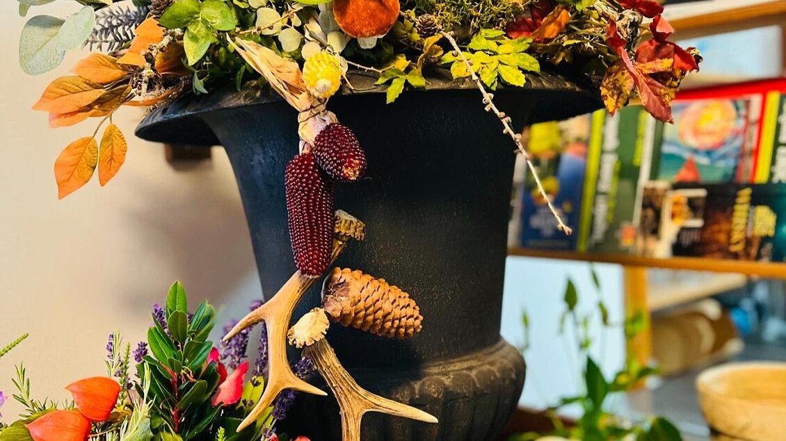 Eine dekorative Blumenvase mit bunten Blumen und herbstlichen Akzenten. Im Hintergrund sind Bücher und weitere Pflanzen sichtbar. | © Wirtshaus Meißl