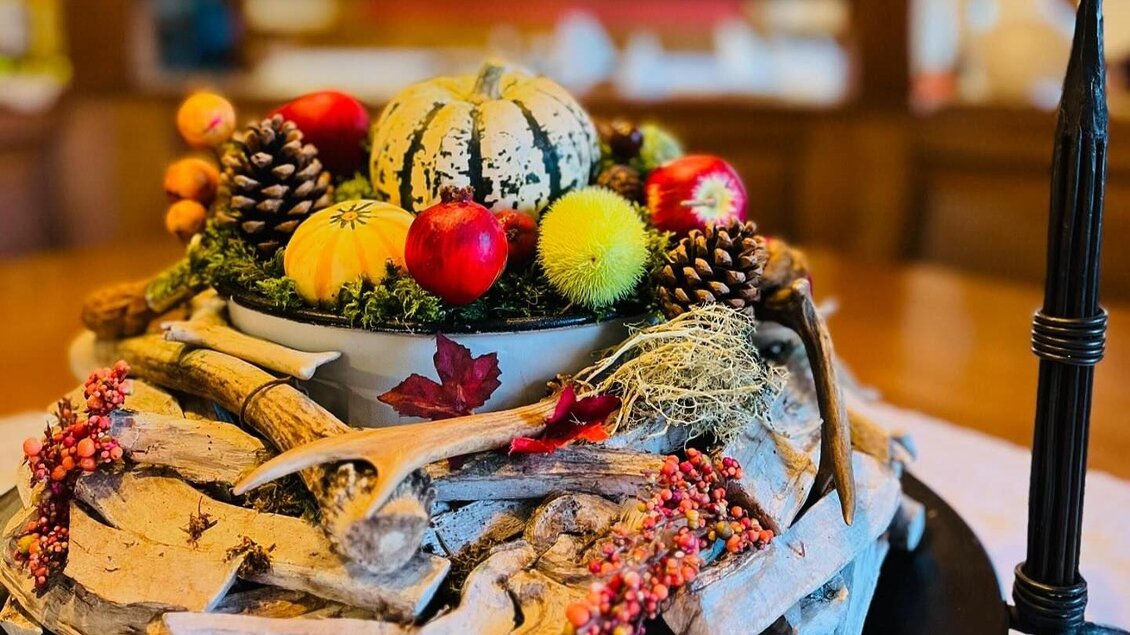 Eine herbstliche Dekoration mit kleinen Kürbissen, Äpfeln und Tannenzapfen in einer Schale. Umgeben von Holzstücken und Beeren, strahlt die Anordnung Gemütlichkeit aus. | © Wirtshaus Meißl