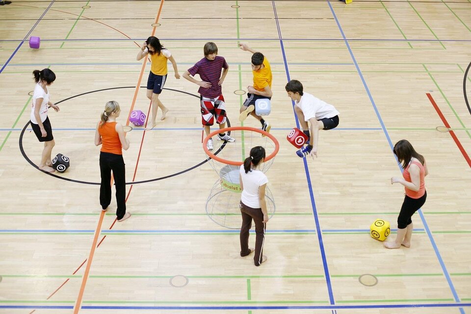 Eine Gruppe von Kindern spielt in einer Sporthalle und bewegt sich aktiv mit bunten Bällen. Einige Kinder stehen in einem Kreis, während andere sich um sie herum bewegen. | © Pixabay