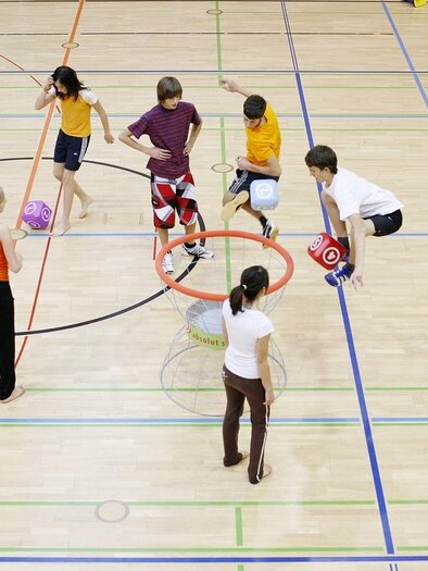 Eine Gruppe von Kindern spielt in einer Sporthalle und bewegt sich aktiv mit bunten Bällen. Einige Kinder stehen in einem Kreis, während andere sich um sie herum bewegen. | © Pixabay