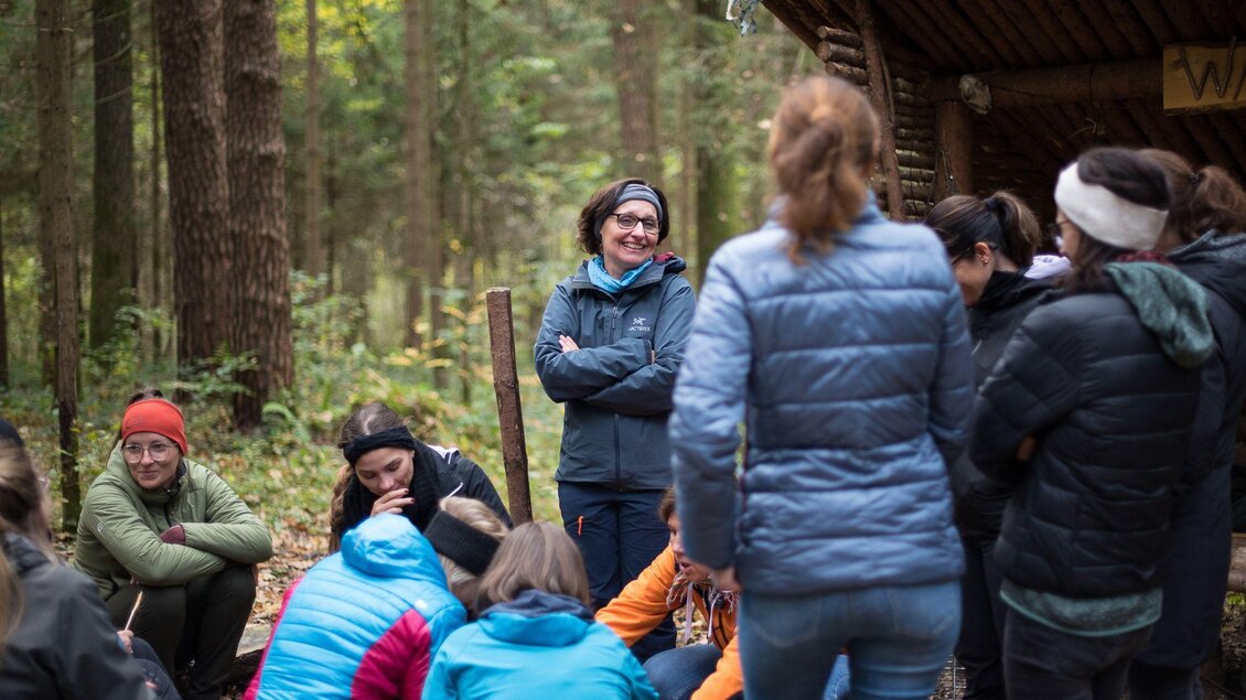 Gruppe Erwachsener im Feriencamp bei der Hirzmannsperre in Edelschrott steht und sitzt im Wald bei einer einfachen Holzhütte. | © Julia C. Pirkenau