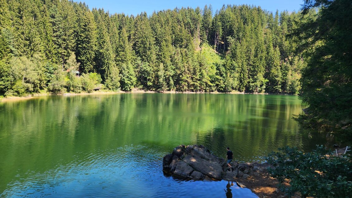 Blick über den Hirzmann-Stausee mit bewaldetem Ufer und einer Person auf einem Felsen. | © Julia C. Pirkenau