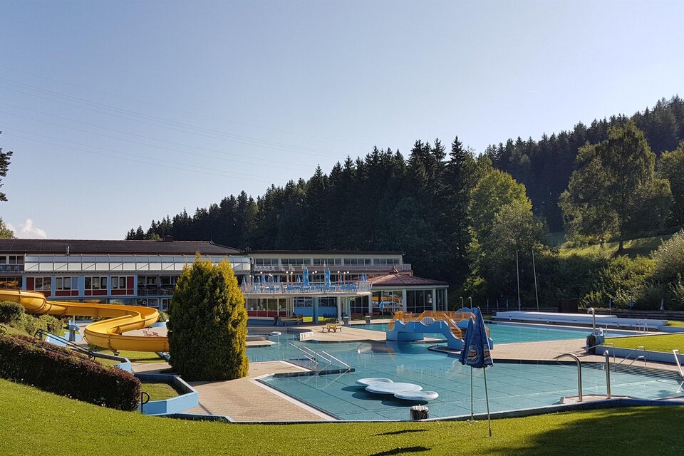 Ein Freibad mit einer Rutsche und mehreren Schwimmbecken umgeben von grünen Wiesen und Bäumen. Im Hintergrund ist ein hellblauer Himmel zu sehen. | © Stadtgemeinde Judenburg