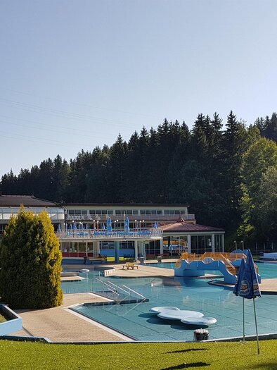 Ein Freibad mit einer Rutsche und mehreren Schwimmbecken umgeben von grünen Wiesen und Bäumen. Im Hintergrund ist ein hellblauer Himmel zu sehen. | © Stadtgemeinde Judenburg