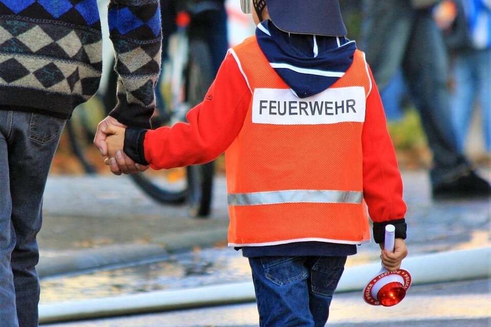 Ein Kind in einem Feuerwehr-Helm und einer leuchtend orangen Weste hält die Hand eines Erwachsenen. Im Hintergrund sind Feuerwehrschläuche zu sehen. | © Pixabay