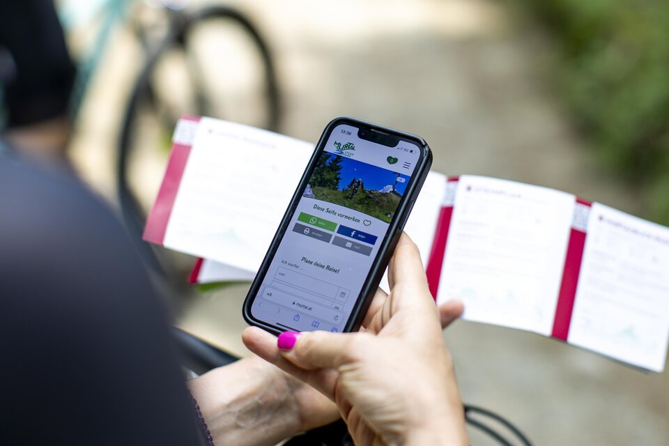 Ein Smartphone, das eine App zeigt, liegt in der Hand. Im Hintergrund sind Informationskarten und ein Fahrrad zu sehen. | © Erlebnisregion Murtal