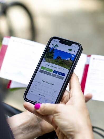Ein Smartphone, das eine App zeigt, liegt in der Hand. Im Hintergrund sind Informationskarten und ein Fahrrad zu sehen. | © Erlebnisregion Murtal