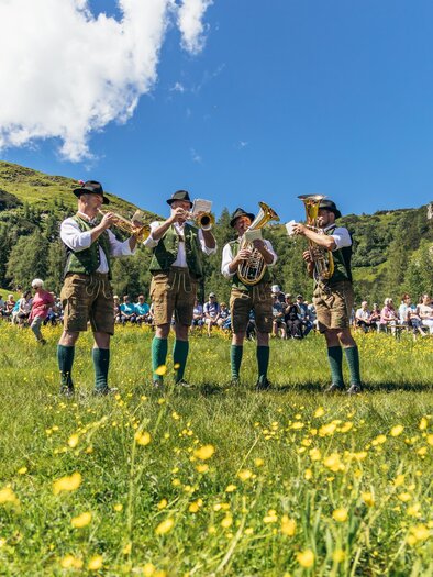 Eine Gruppe von vier Musikern spielt traditionelle Blasmusik in einer grünen Wiese. Im Hintergrund sind viele Zuschauer und die malerische Berglandschaft zu sehen. | © Reiteralm Bergbahnen GmbH & Co KG