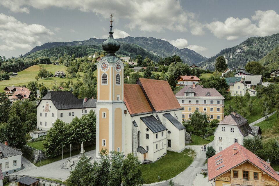 Eine malerische Dorflandschaft mit einer auffälligen Kirche und umliegenden Bergen. Die Gebäude sind bunt und harmonisch in die Umgebung eingegliedert. | © WEGES