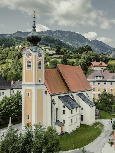 Eine malerische Dorflandschaft mit einer auffälligen Kirche und umliegenden Bergen. Die Gebäude sind bunt und harmonisch in die Umgebung eingegliedert. | © WEGES