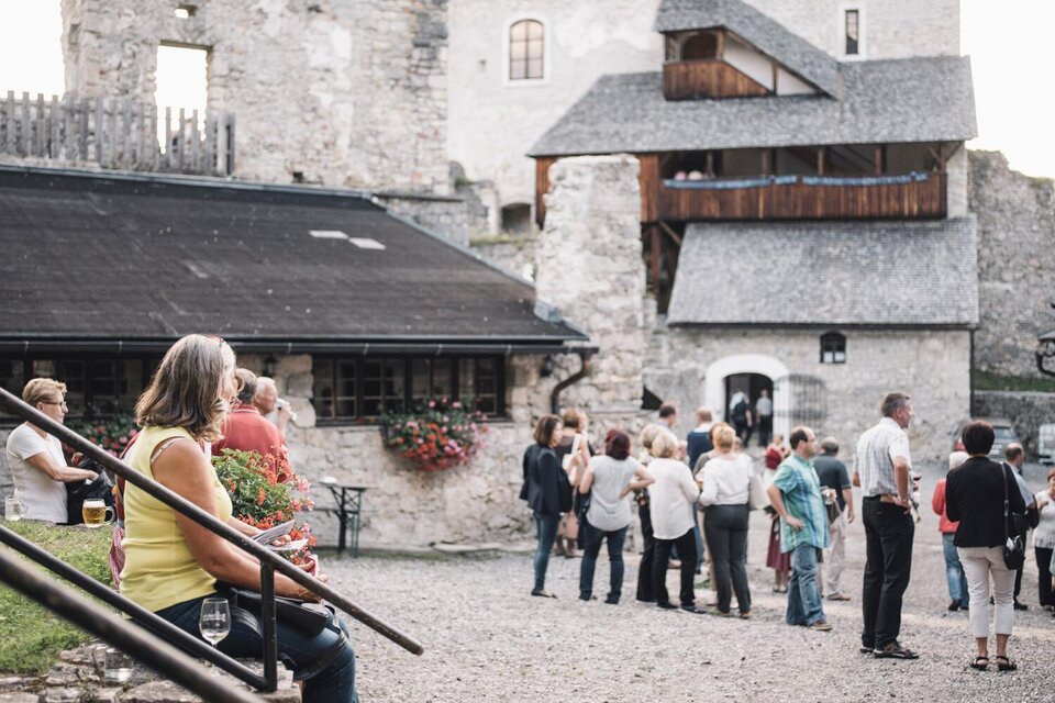 Eine Gruppe von Menschen versammelt sich auf einem Platz vor einer historischen Burg. Im Vordergrund sitzt eine Frau auf einer Treppe und hält einen Pflanzentopf. | © Stefan Leitner