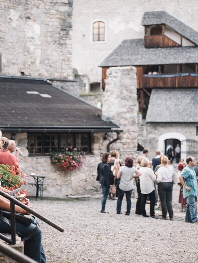 Eine Gruppe von Menschen versammelt sich auf einem Platz vor einer historischen Burg. Im Vordergrund sitzt eine Frau auf einer Treppe und hält einen Pflanzentopf. | © Stefan Leitner