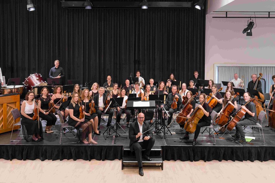 Ein großes Orchester steht auf einer Bühne und spielt Musik. Im Vordergrund sitzt der Dirigent und leitet die Musiker. | © Foto Schleich