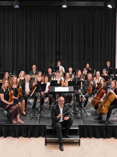 Ein großes Orchester steht auf einer Bühne und spielt Musik. Im Vordergrund sitzt der Dirigent und leitet die Musiker. | © Foto Schleich