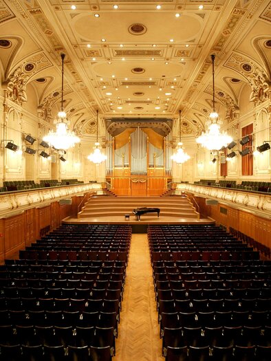 An elegant concert hall with many seats and a large organ in the background. The ceiling is designed with impressive light fixtures and decorative elements. | © Ilemann
