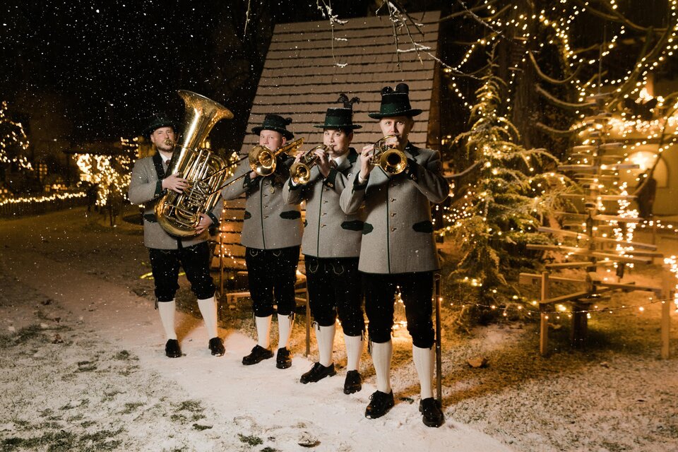 Vier Musiker in traditioneller Tracht spielen im Schnee. Im Hintergrund leuchten festliche Lichter und es schneit leicht. | © Lupi Spuma