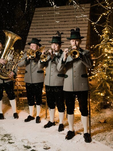 Four musicians in traditional attire are playing in the snow. In the background, festive lights are glowing and it is lightly snowing. | © Lupi Spuma