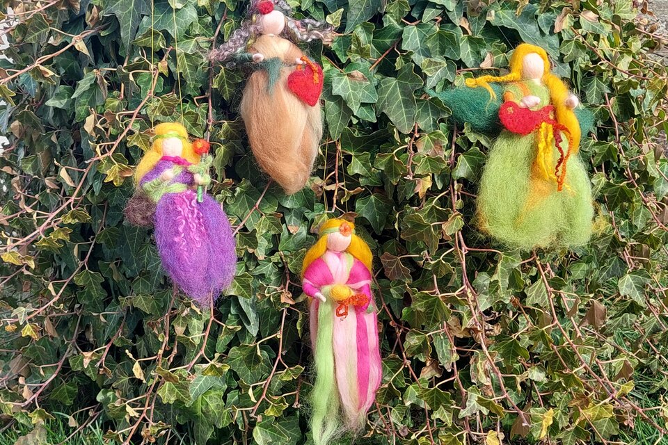 Vier handgemachte Figuren hängen an einer grünen Efeuwand. Jede Figur hat ein einzigartiges Design und farbige Kleider.