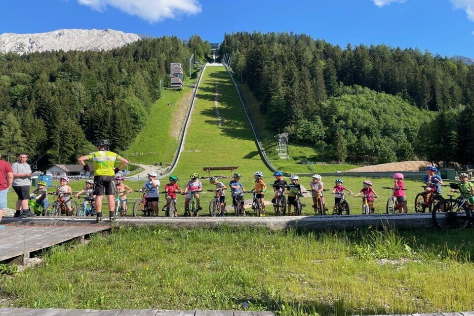Die zahlreichen Kinder beim Kinder Radcup am Kulm warten geduldig auf ihren Start. Sie sitzen nebeneinander aufgereiht auf ihren Fahrrädern. Im Hintergrund ist der Kulm mit seiner berühmten Skiflugschanze zu sehen, die im sommerlichen grün erstrahlt. Sie ist umgeben von grünen Bäumen, und im Hintergrund ist der mächtige Hausberg des Hinterberger Tals, der Grimming, zu sehen. | © Auszeit Ausseerland