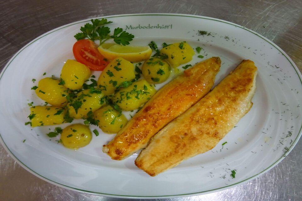 Ein köstlicher Teller mit gebratenem Fisch, begleitet von Kartoffeln und frischen Kräutern. Eine Zitronenscheibe und eine Tomate sorgen für zusätzliche Frische. | © Gasthaus Murbodenhüttl