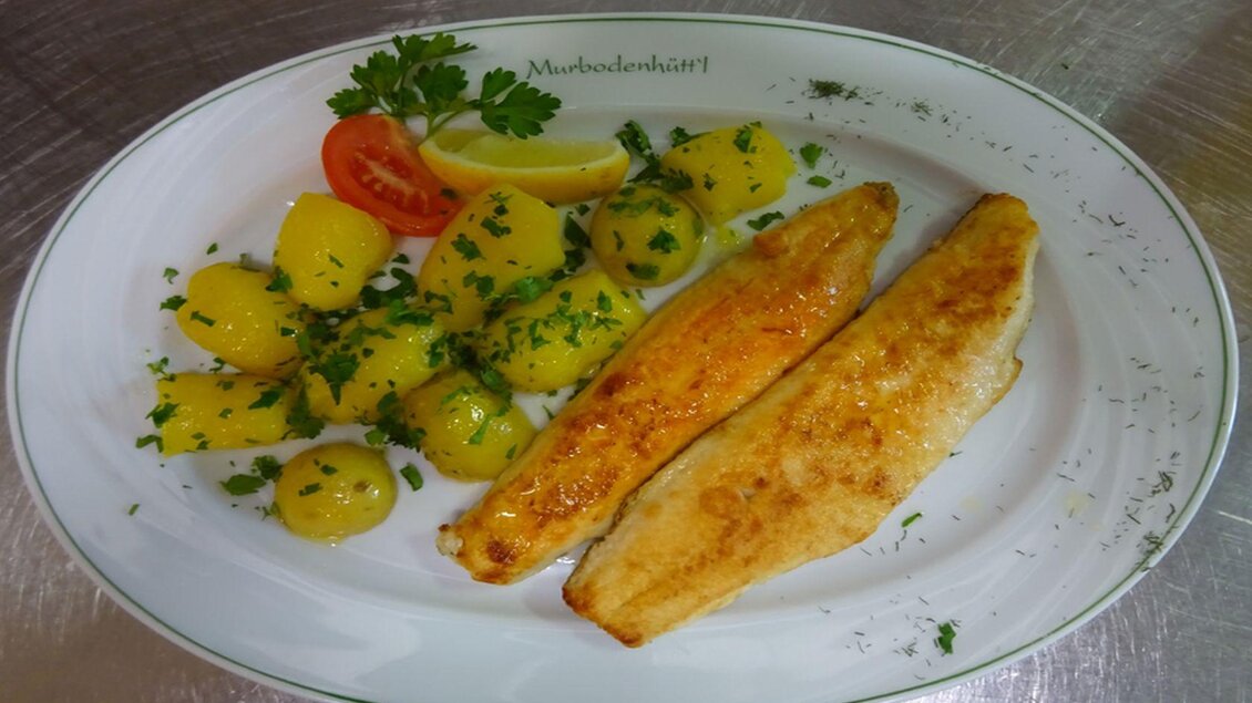 Ein köstlicher Teller mit gebratenem Fisch, begleitet von Kartoffeln und frischen Kräutern. Eine Zitronenscheibe und eine Tomate sorgen für zusätzliche Frische. | © Gasthaus Murbodenhüttl