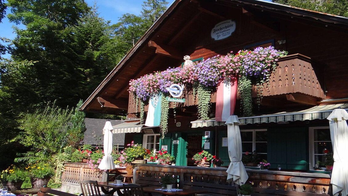 Ein charmantes, rustikales Gasthaus umgeben von Bäumen und bunten Blumen. Der Balkon ist mit Pflanzen dekoriert, und Tische stehen im Freien zum Verweilen bereit. | © Fischerhütte/Syen
