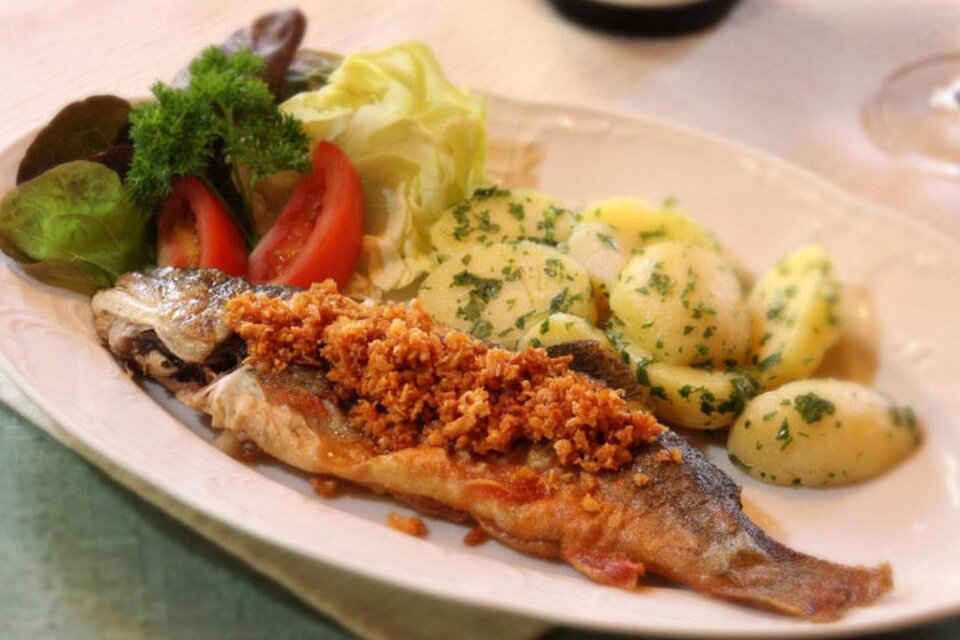 Ein köstlicher gebratener Fisch mit knuspriger Kruste, serviert mit Kartoffeln und frischem Salat. Auf dem Teller sind auch Tomaten und grüne Blätter zu sehen. | © Syen Andreas