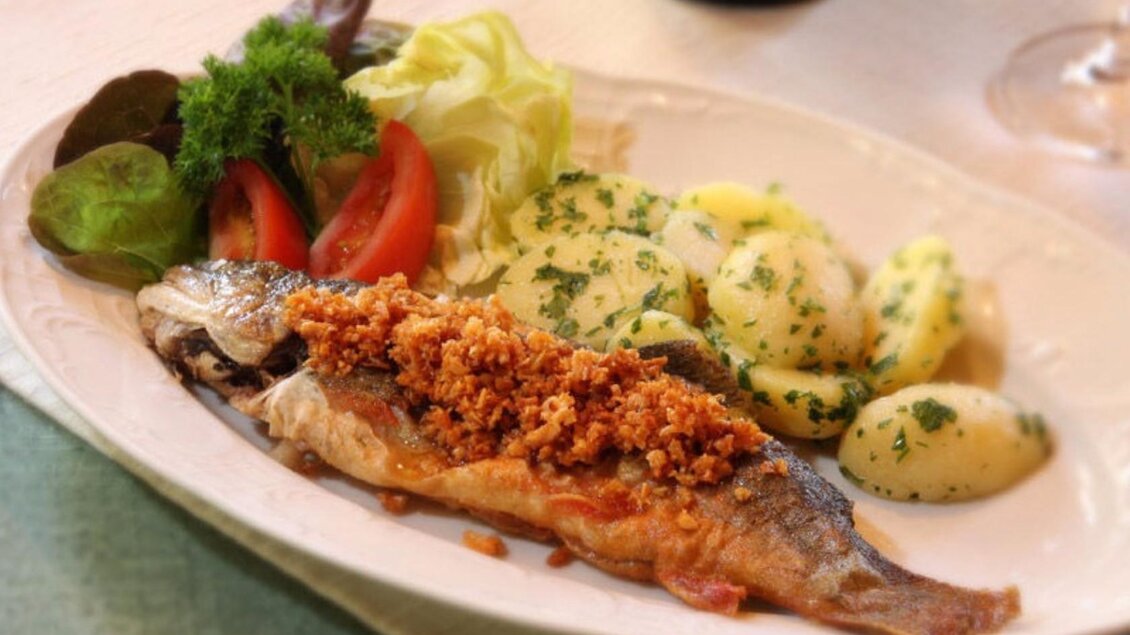 Ein köstlicher gebratener Fisch mit knuspriger Kruste, serviert mit Kartoffeln und frischem Salat. Auf dem Teller sind auch Tomaten und grüne Blätter zu sehen. | © Syen Andreas